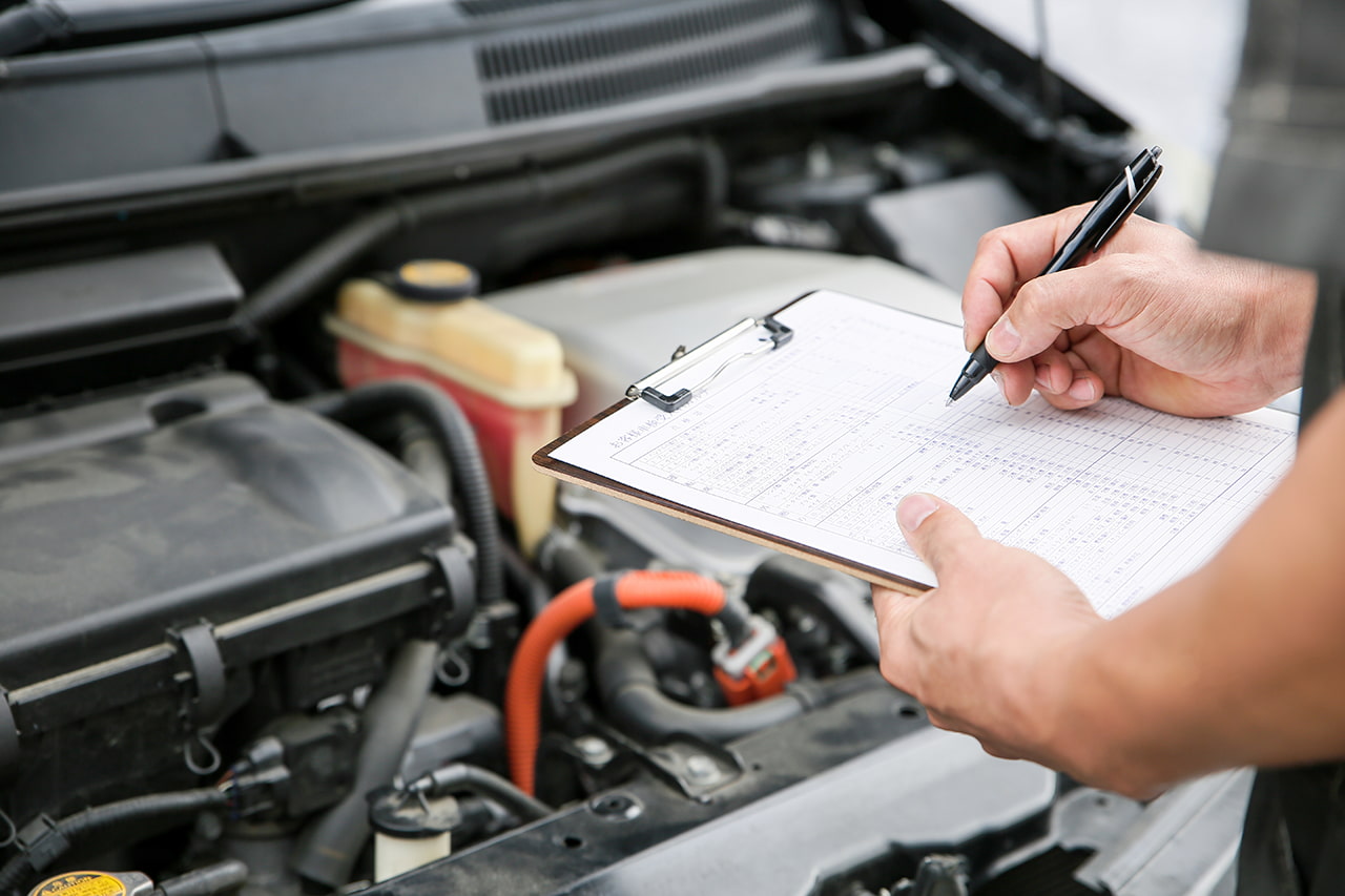 車のエンジンルームを前に、バインダーに挟んだ書類へ記入する手元